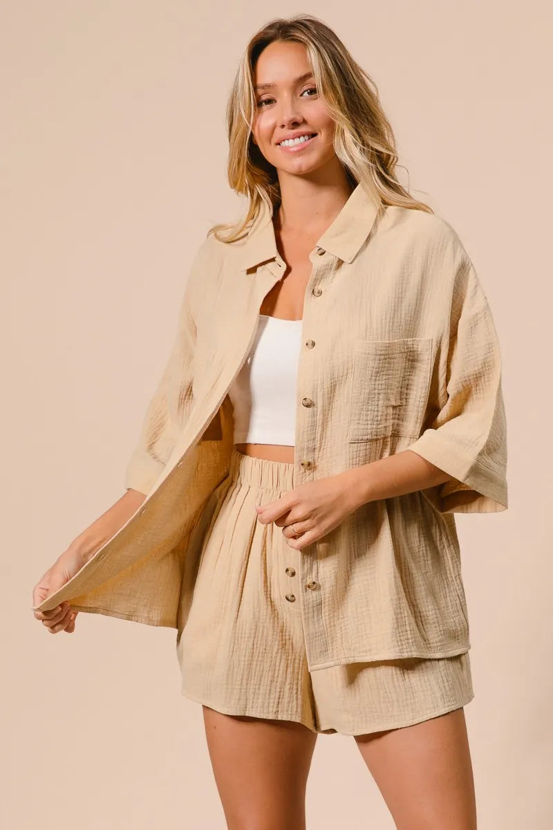 Woman in beige linen button-up shirt and matching shorts over white crop top, smiling against neutral background in relaxed pose.