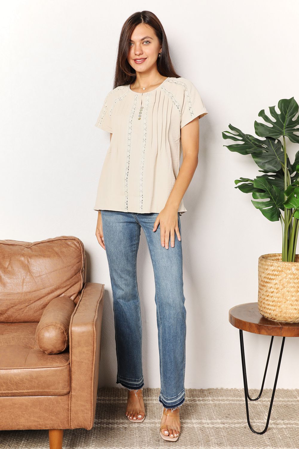 Woman wearing beige crochet buttoned short sleeves top and jeans standing in a chic living room with stylish decor.