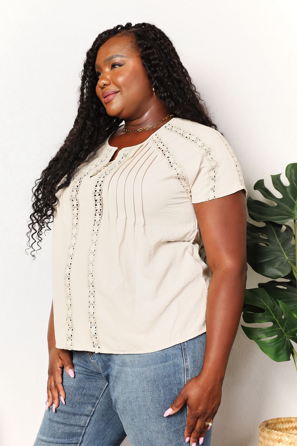 Woman wearing a beige crochet buttoned short sleeves top with blue jeans, standing near a green plant.