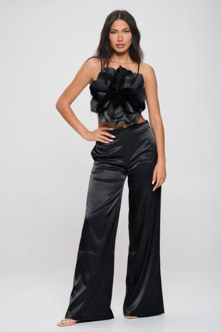 Woman in black satin two-piece outfit with dramatic ruffled strapless top and wide-leg pants, posed with hand on hip against light gray background.