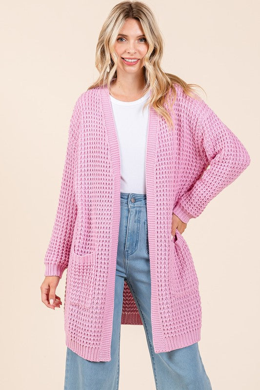 Woman wearing a pink waffle-knit open front longline cardigan with pockets, styled casually with a white top and jeans.