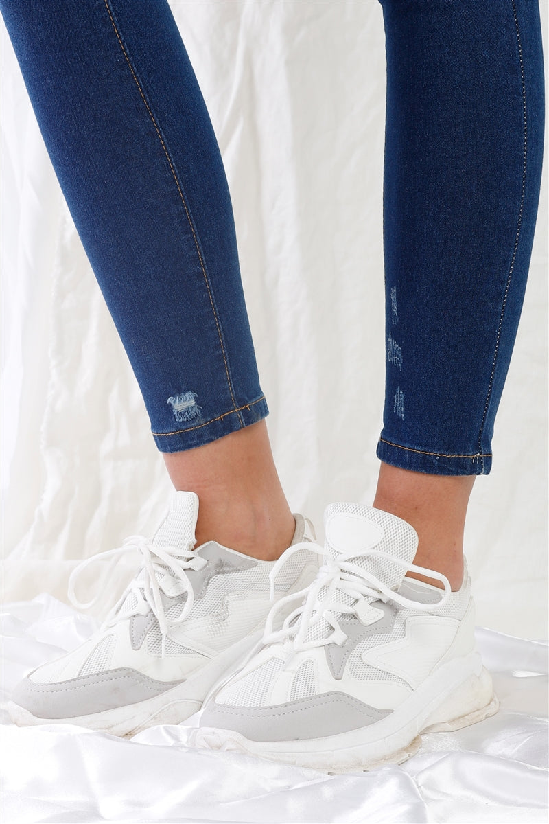 Dark blue high-waisted skinny jeans with small rips near hem, paired with white sneakers, showcasing casual and stylish look.