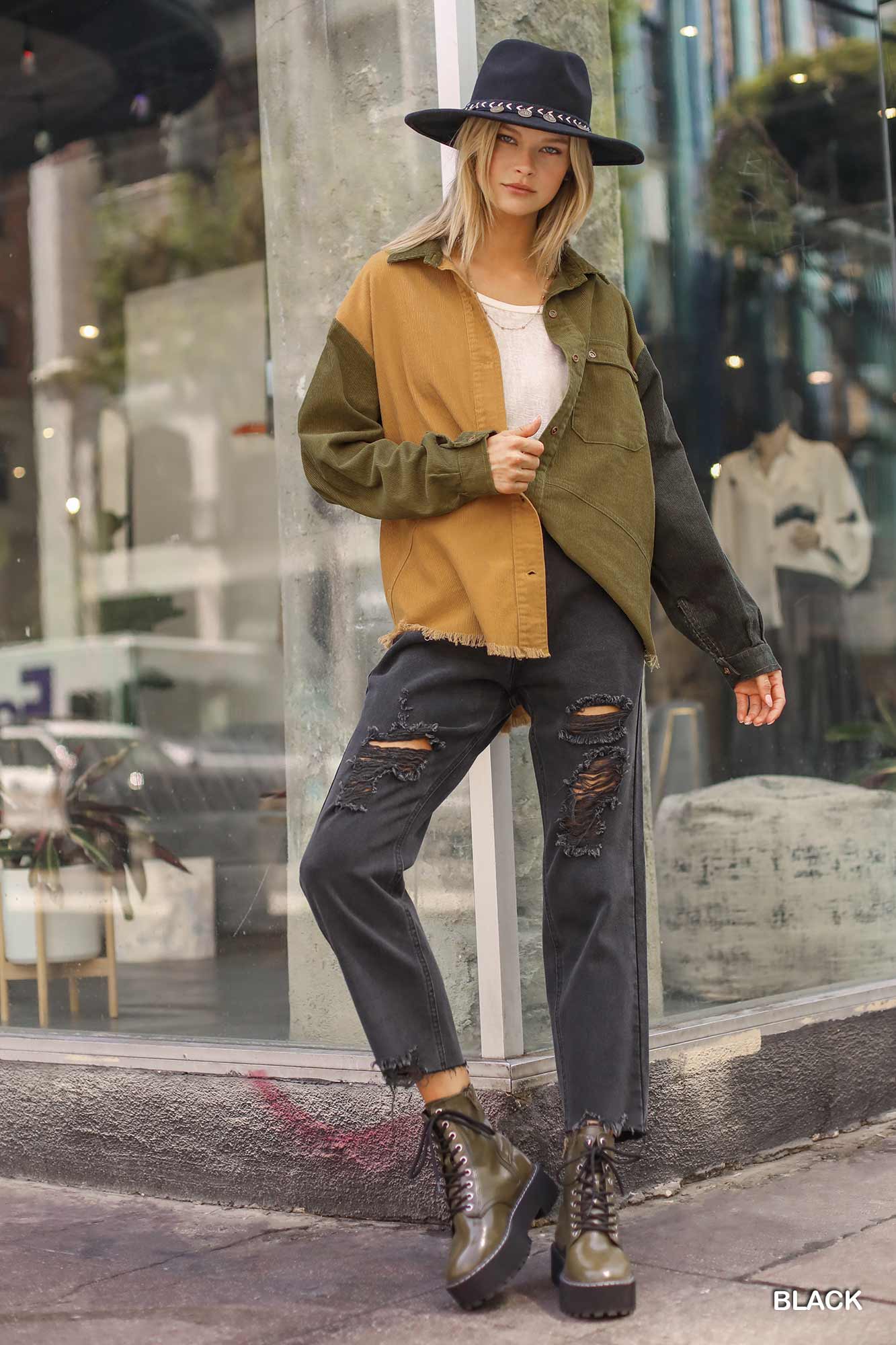 Woman wearing straight cut distressed black denim jeans with raw hem, styled with a two-tone jacket and hat, standing on a city sidewalk.