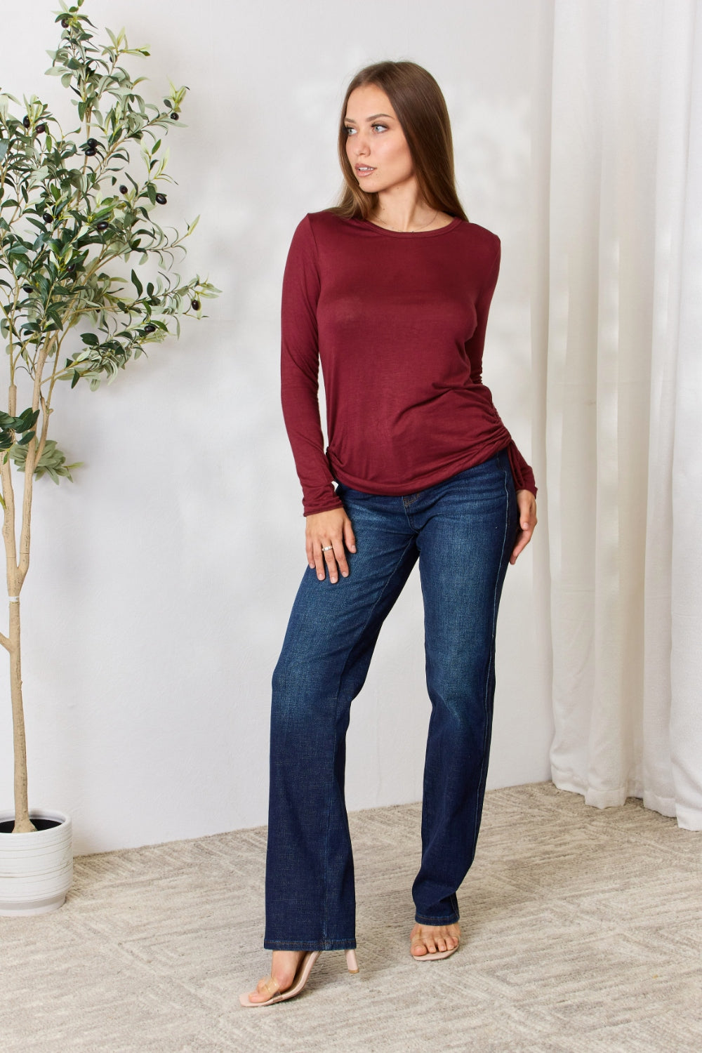 Woman wearing burgundy drawstring round neck long sleeve top with dark jeans, standing in a stylish room with a plant.