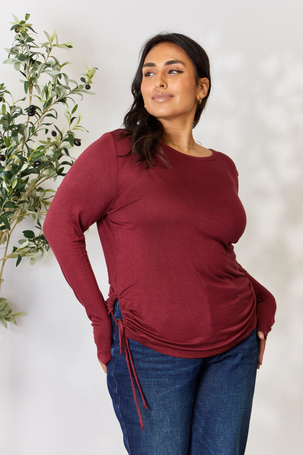 Woman wearing a drawstring round neck long sleeve top in burgundy, paired with blue jeans, standing near a plant.