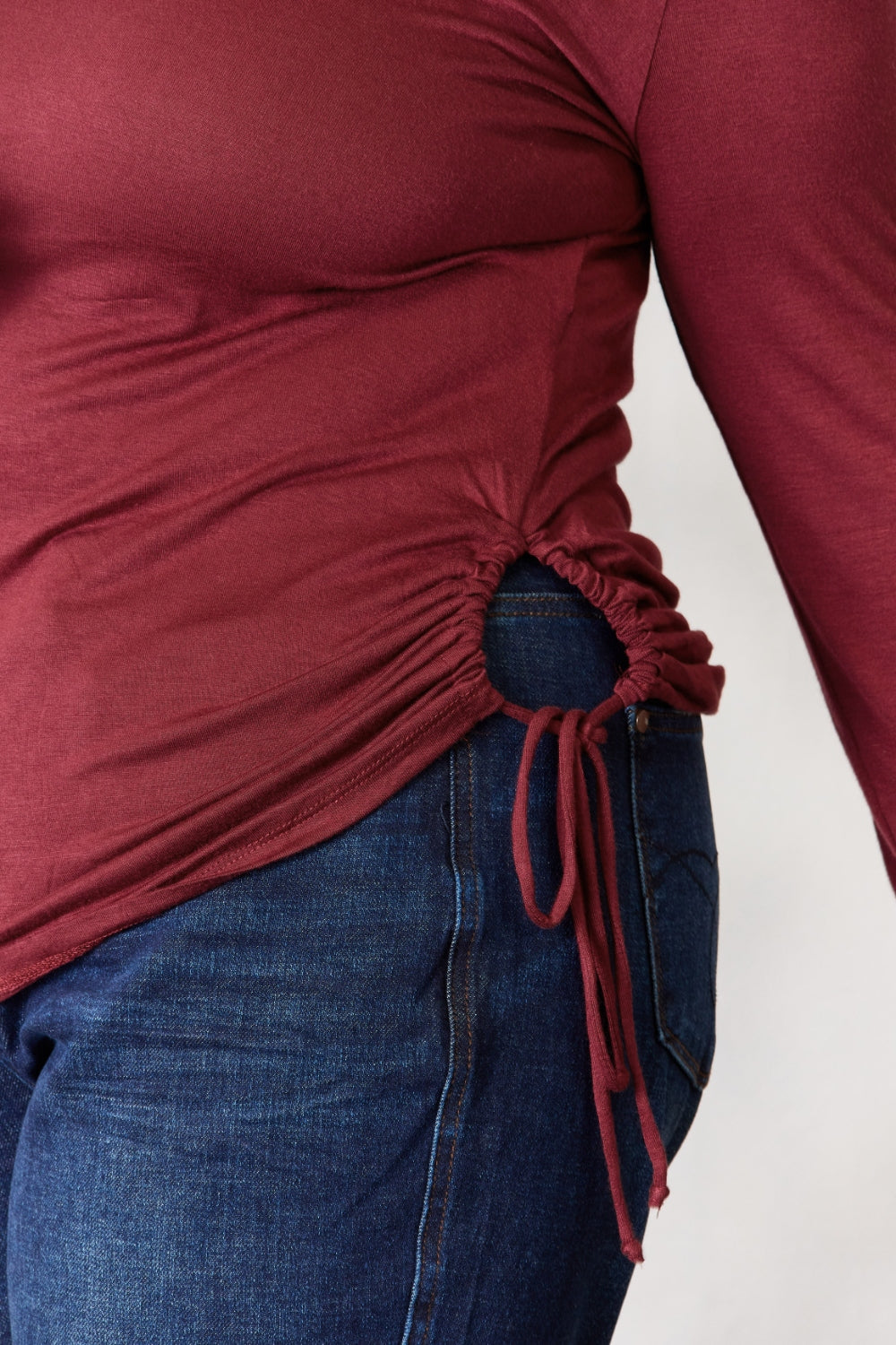 Burgundy drawstring round neck long sleeve top, showing side detail with adjustable tie, paired with blue jeans.