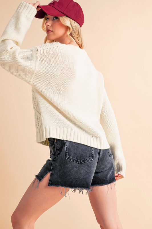 Woman wearing white cable knit drop shoulder sweater with denim shorts and a red cap, showcasing casual fashion style.