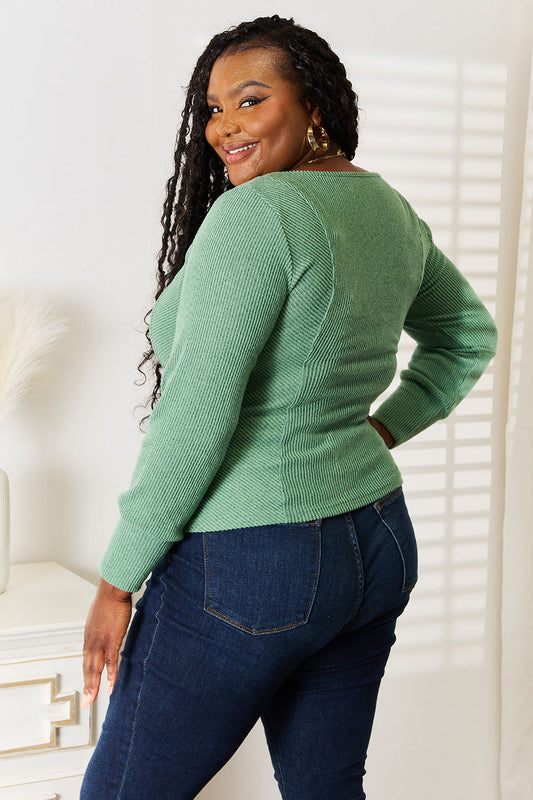 Woman wearing green scoop neck long sleeve top with blue jeans, standing sideways while smiling in a softly lit room.