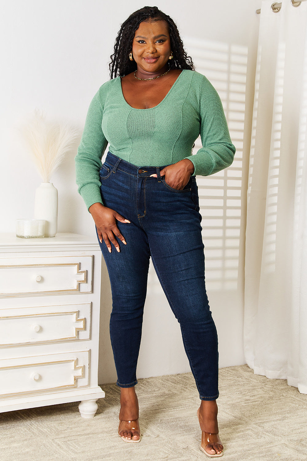 Woman wearing green scoop neck long sleeve top and jeans, standing next to a white dresser in a bright room.