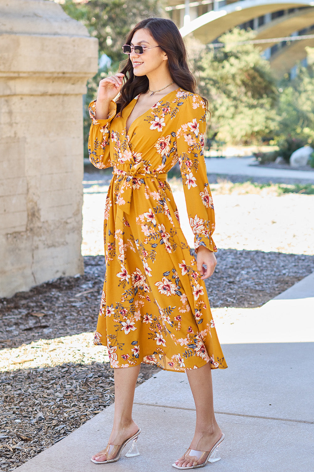 Woman wearing a floral tie-back flounce sleeve dress in mustard yellow with pink and white flowers, standing outdoors.