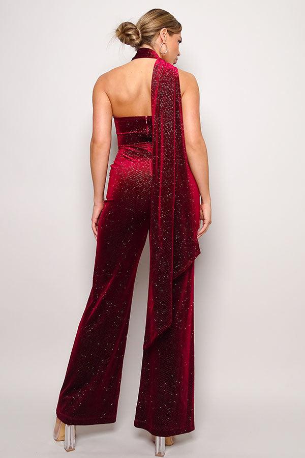 Woman wearing sleeveless red glitter velvet jumpsuit with one-shoulder design, open back, and long pants with scarf detail.