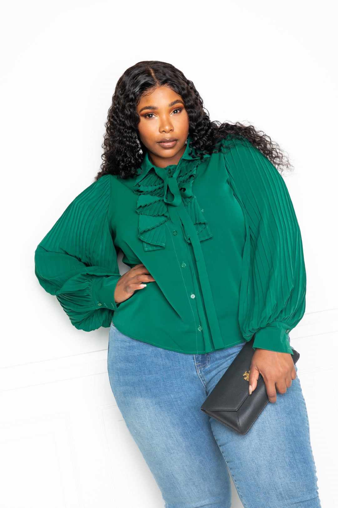 Plus-size model in green pleated sleeve blouse with bow and frill, paired with jeans and holding a clutch.