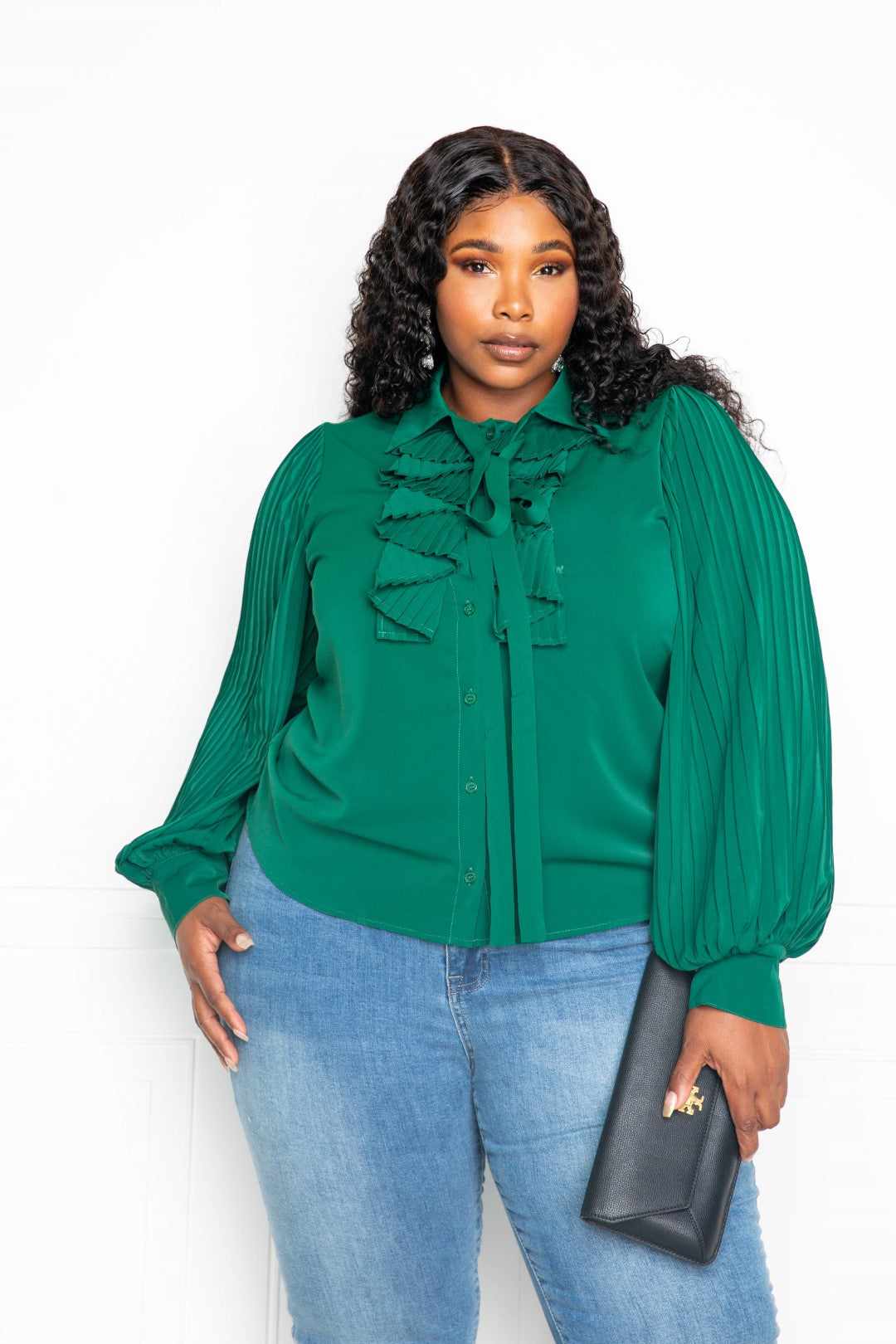 Woman wearing 1XL forrest green pleated sleeve blouse with waterfall frill and bow, paired with jeans, holding a black clutch.