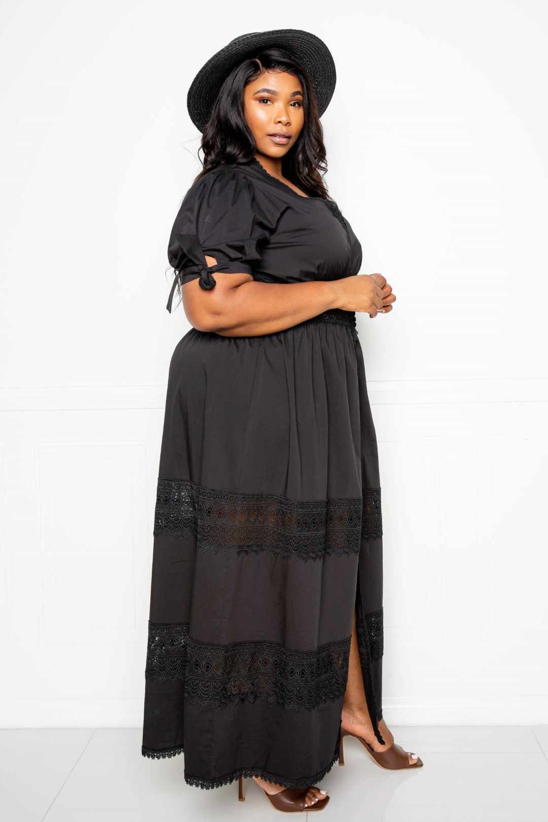 Plus size model wearing a black puff sleeve maxi dress with lace insert, styled with a black hat and heels. Perfect for elegant occasions.