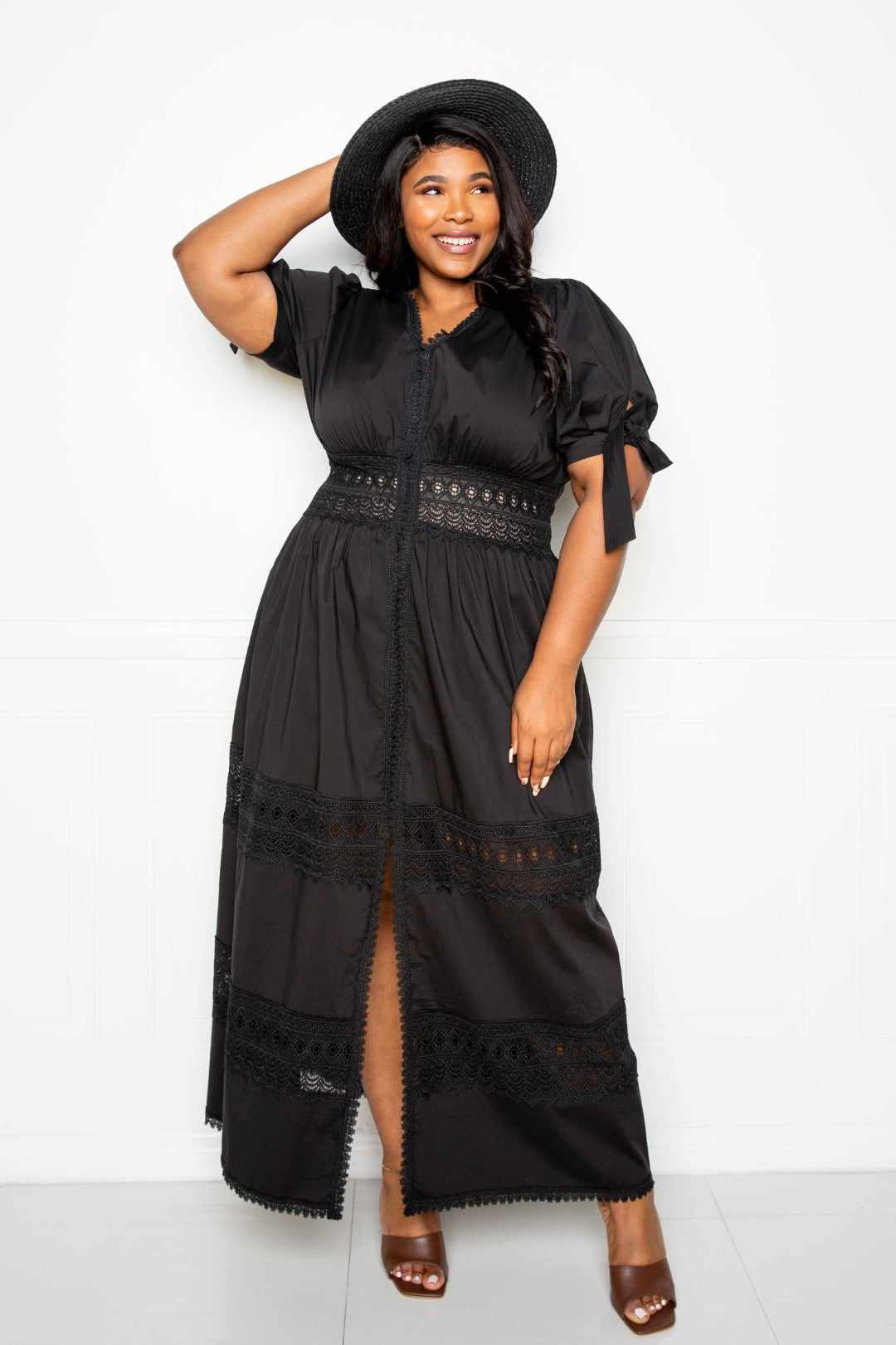 Woman wearing black puff sleeve maxi dress with lace insert and hat, smiling, hand on head, styled with open toe heels.