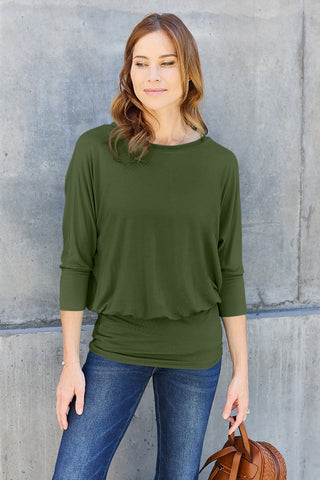 Woman wearing green round neck batwing sleeve top, paired with blue jeans, standing against a concrete wall.