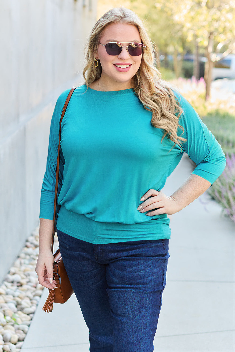 Woman wearing teal round neck batwing sleeve top with sunglasses, styled with jeans outdoors.