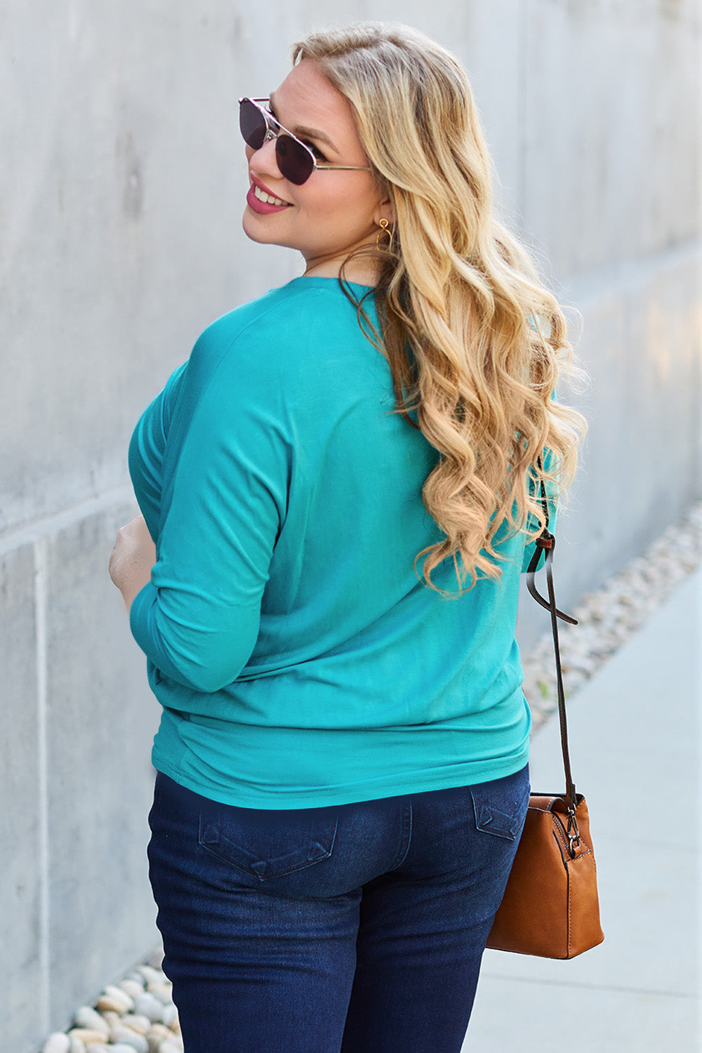 Woman wearing a turquoise round neck batwing sleeve top with sunglasses and jeans, carrying a brown purse, outdoors.