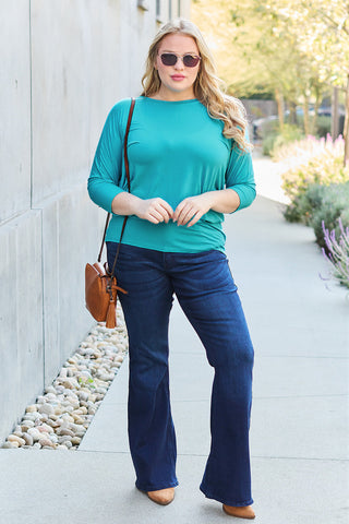 Woman wearing teal round neck batwing sleeve top with jeans, accessorized with sunglasses and a brown shoulder bag, standing outdoors.