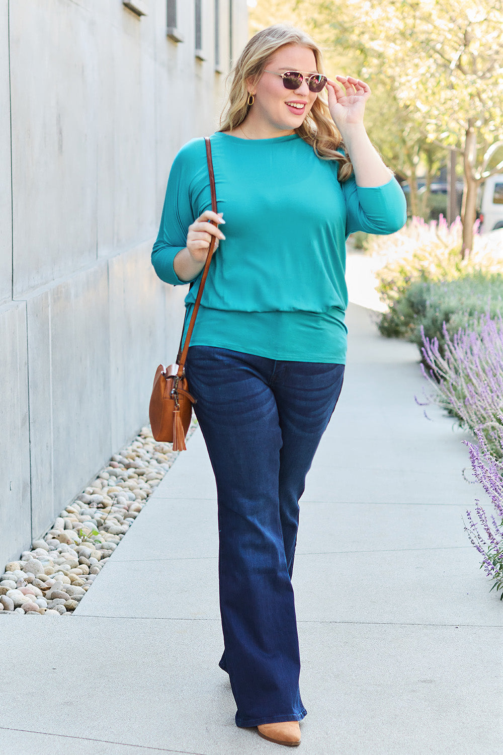 Woman wearing round neck batwing sleeve top in teal with jeans and sunglasses, walking outdoors on a sunny day.