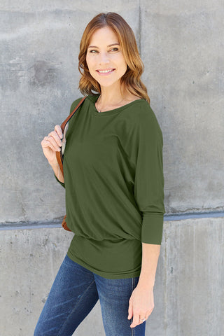 Woman wearing a round neck batwing sleeve top, made of 95% rayon and 5% spandex, standing against a concrete wall.