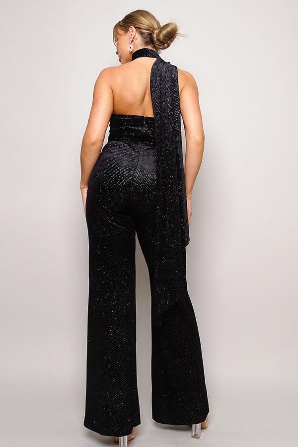 Woman wearing a glitter velvet jumpsuit with scarf detail, featuring an open back and wide-leg silhouette for an elegant look.