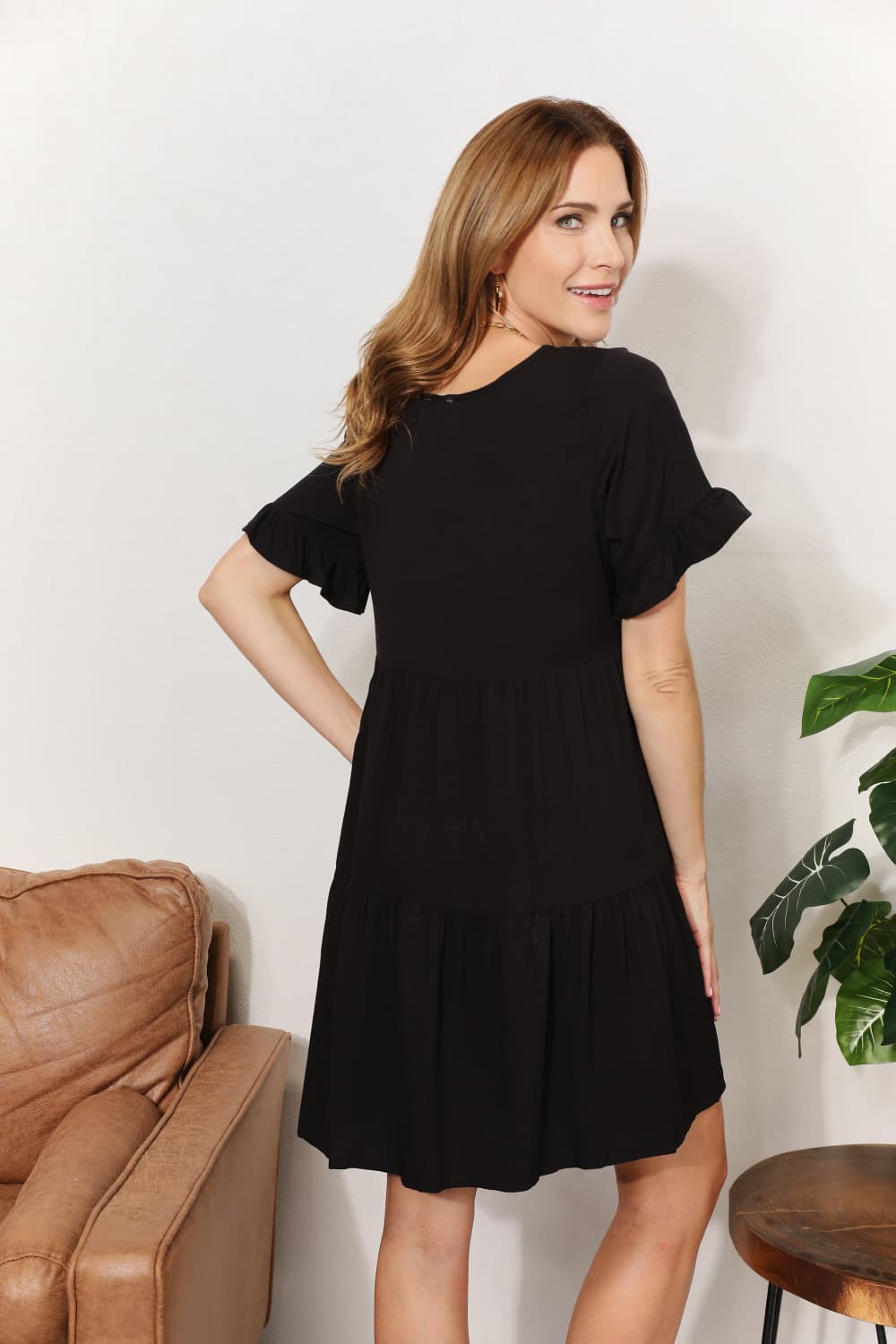 Woman wearing black V-neck flounce sleeve tiered dress with A-line hem, standing in stylish room with plants and brown chair.