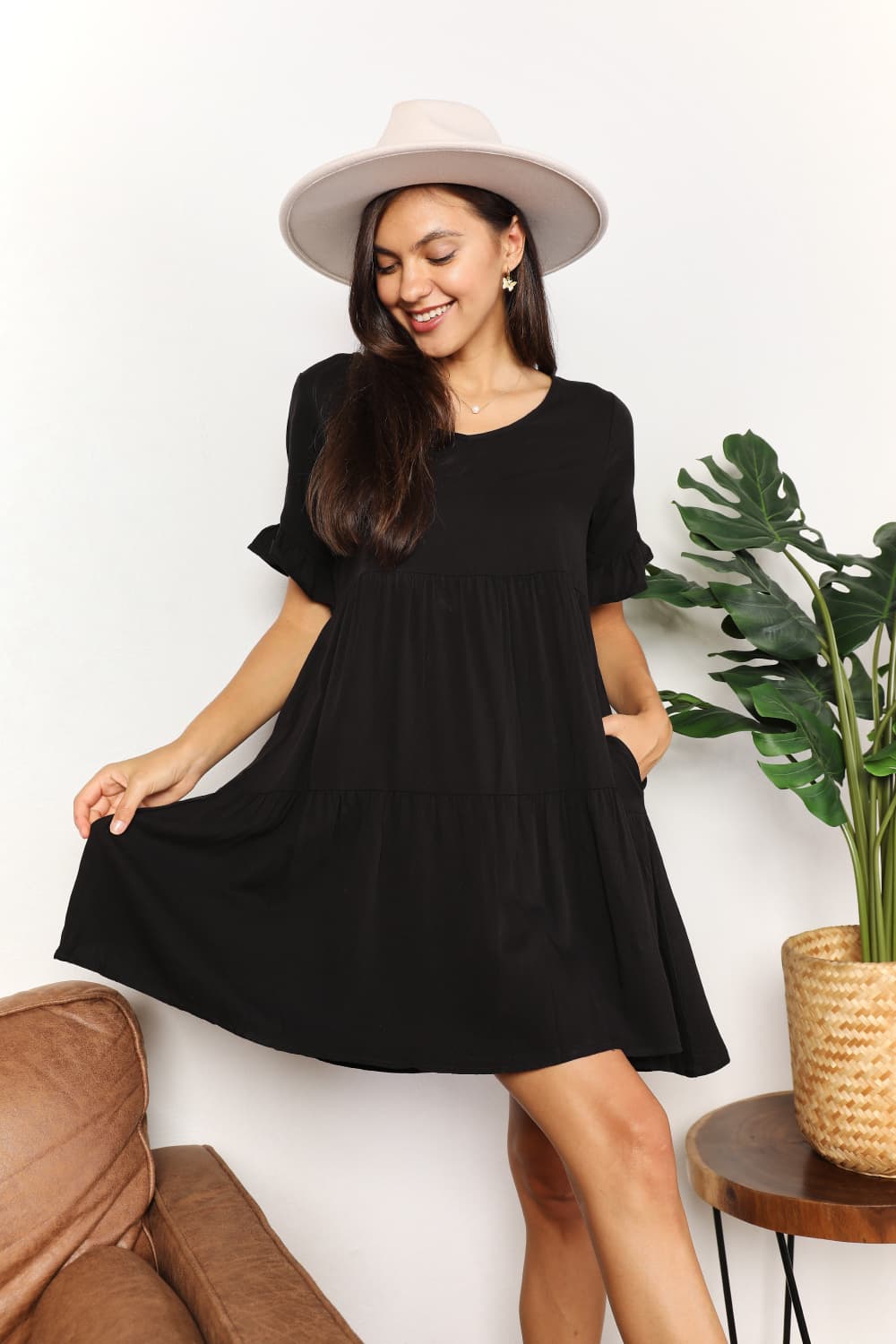 Woman wearing a black V-neck flounce sleeve tiered dress with a stylish hat, standing indoors near a plant and table.