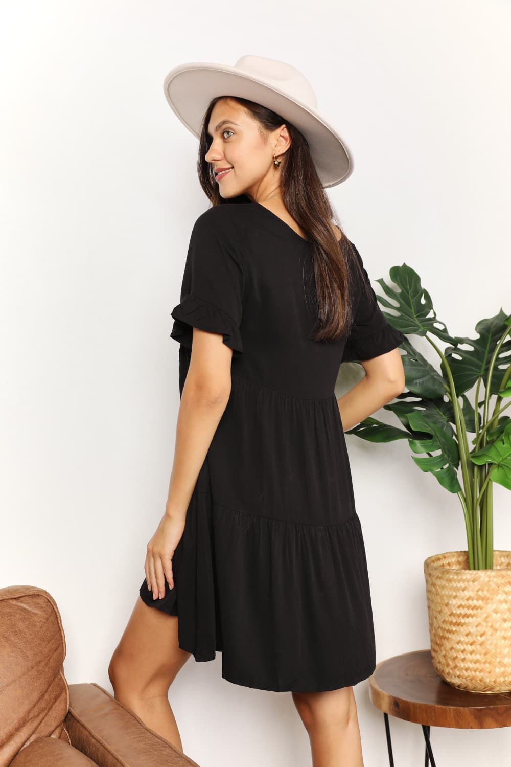 Woman wearing a black V-neck flounce sleeve tiered dress with a wide-brim hat, standing by a potted plant.