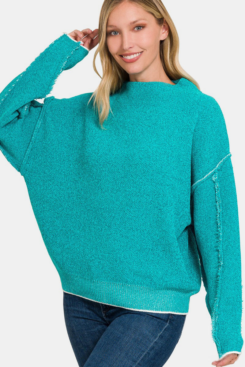 Woman wearing a stylish turquoise mock neck sweater with exposed seam details and long sleeves, paired with blue jeans.