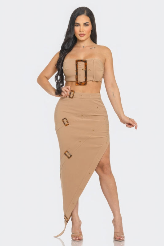 Woman in tan/beige two-piece outfit with tortoiseshell buckle details, featuring a strapless crop top and high-waisted asymmetrical skirt with side...