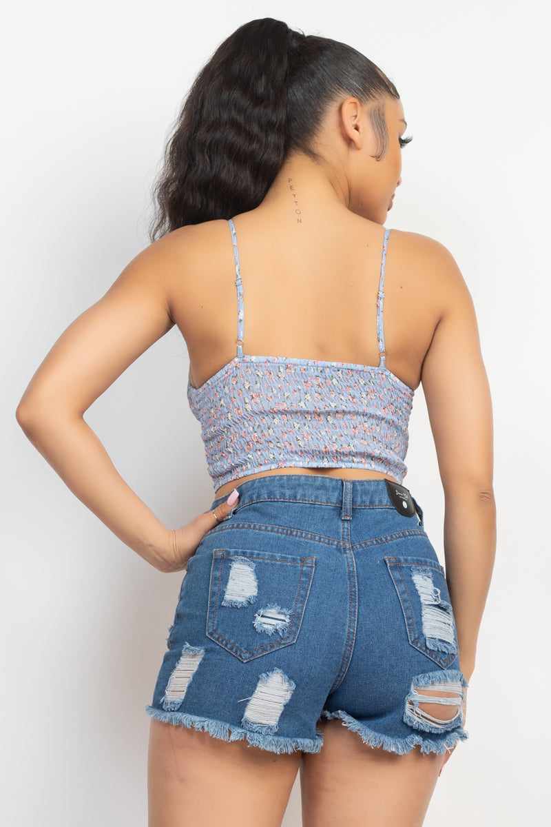 Woman wearing blue ditsy floral front tie top with smocked back and adjustable straps, paired with ripped denim shorts, showcasing back view.