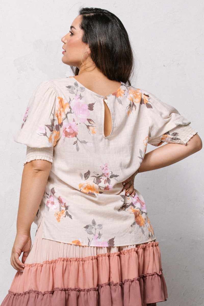 Woman wearing floral printed woven blouse with back button closure, short puffed sleeves, and smocked hem in oatmeal color.