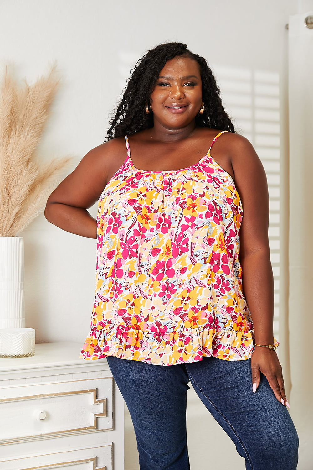 Woman in Double Take Floral Scoop Neck Ruffle Hem Cami with vibrant pattern and ruffle detailing, styled with blue jeans.