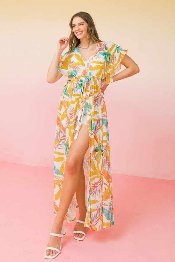 Woman wearing a colorful printed woven maxi cover up with an open front and tie at empire, featuring ruffled sleeves, standing on a pink background.