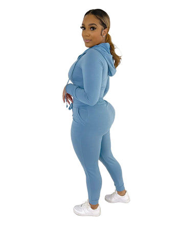 Woman wearing blue French Terry Jacket & Pants Set with hood, side profile, standing indoors against a white paneled wall.