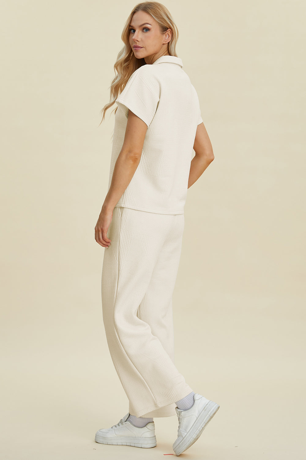 Woman wearing full size collared neck short sleeve top and pants set, two-piece in cream, slightly stretchy fabric, side view.