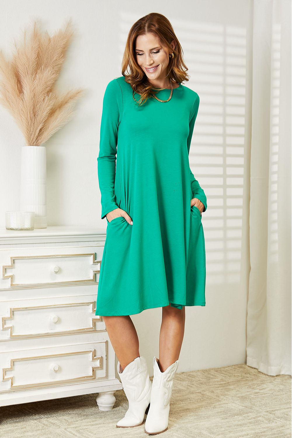 Woman wearing full-size long sleeve green flare dress with pockets, styled with white boots, standing in a modern room.