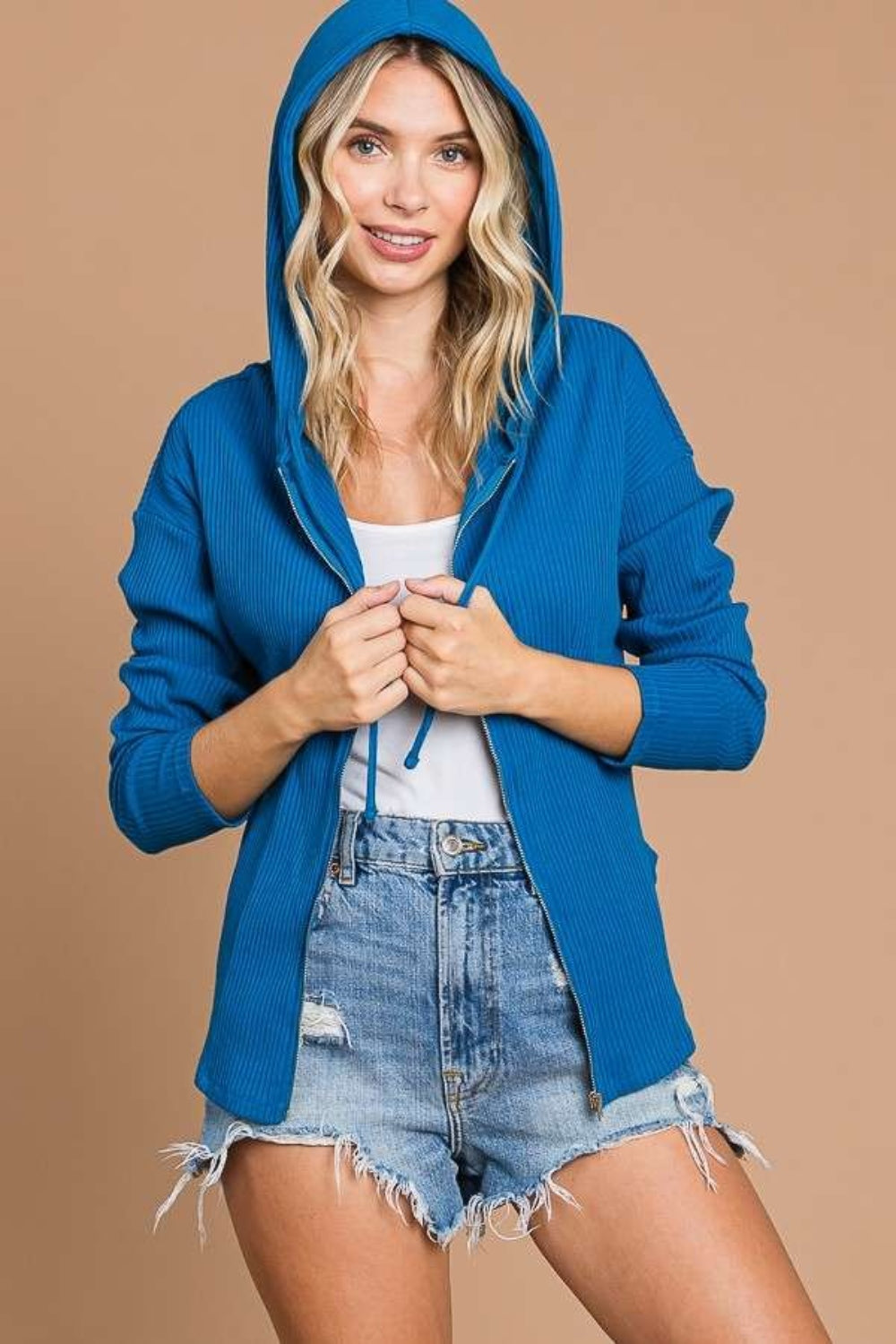 Woman wearing blue ribbed drawstring hooded jacket over white top and denim shorts, styled for a casual look.