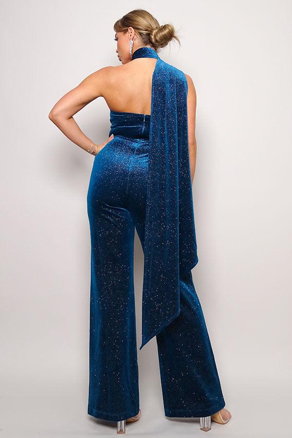 Woman wearing teal glitter velvet jumpsuit with scarf detail, one-shoulder design, and open back.