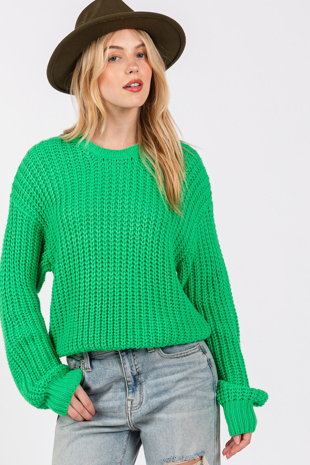 Woman wearing a green round neck drop shoulder sweater made of 100% acrylic, paired with ripped jeans and a stylish hat.