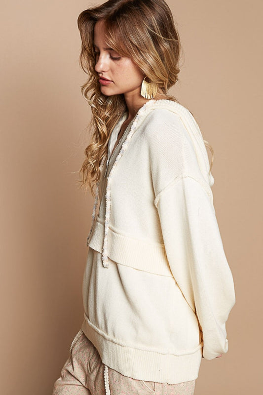 Woman wearing a cream half zip drop shoulder hooded sweater with drawstring and ribbed details, made of acrylic and cotton blend.