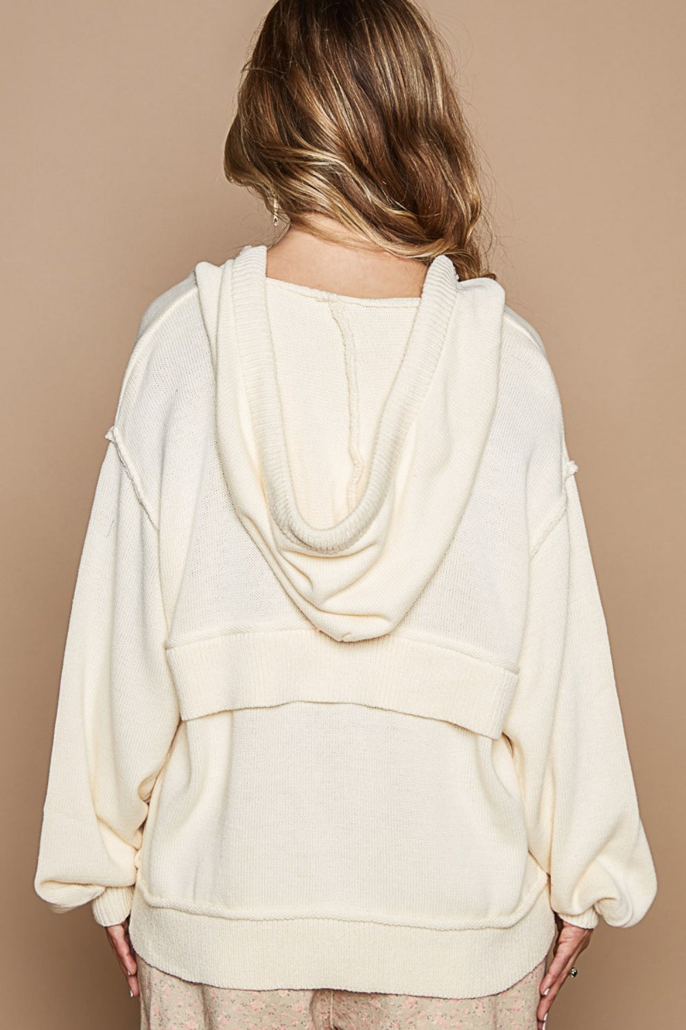 Woman wearing a cream half zip drop shoulder hooded sweater with drawstring and ribbed details, made of 55% acrylic and 45% cotton.