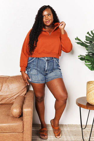 Woman in chic half-zip long sleeve hoodie with raglan sleeves and denim shorts, standing by a couch and table with plant.