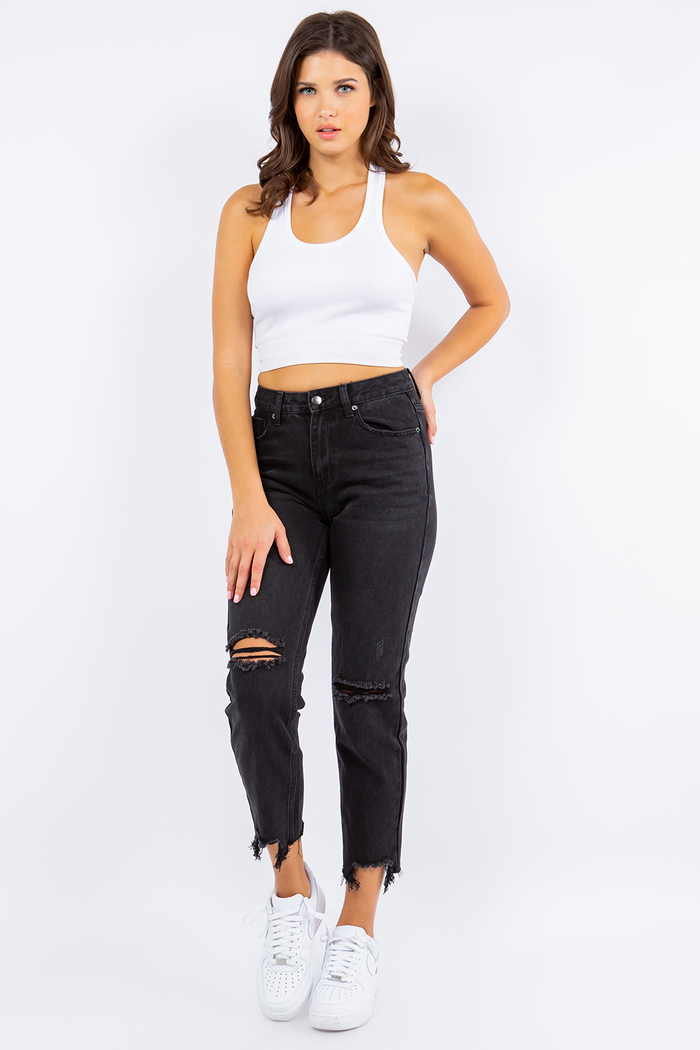 Woman wearing high waist distressed cropped straight jeans with a white tank top and sneakers, showcasing trendy denim style.