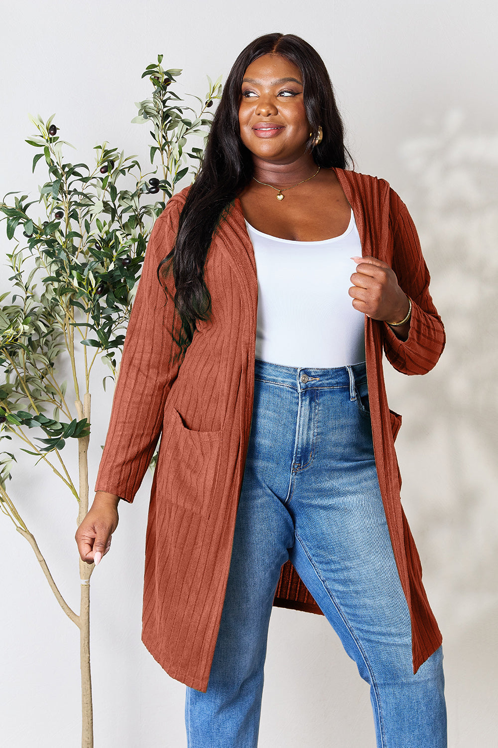 Woman wearing a rust-colored hooded sweater cardigan with pockets, paired with a white top and blue jeans, standing by a plant.