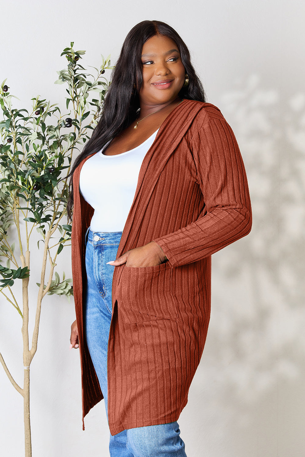 Woman wearing a brown hooded sweater cardigan with pockets, paired with a white top and jeans, standing next to a plant.