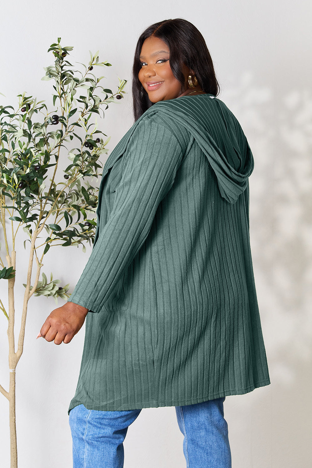 Woman wearing a green, slightly stretchy hooded sweater cardigan with pockets, standing next to an indoor plant.