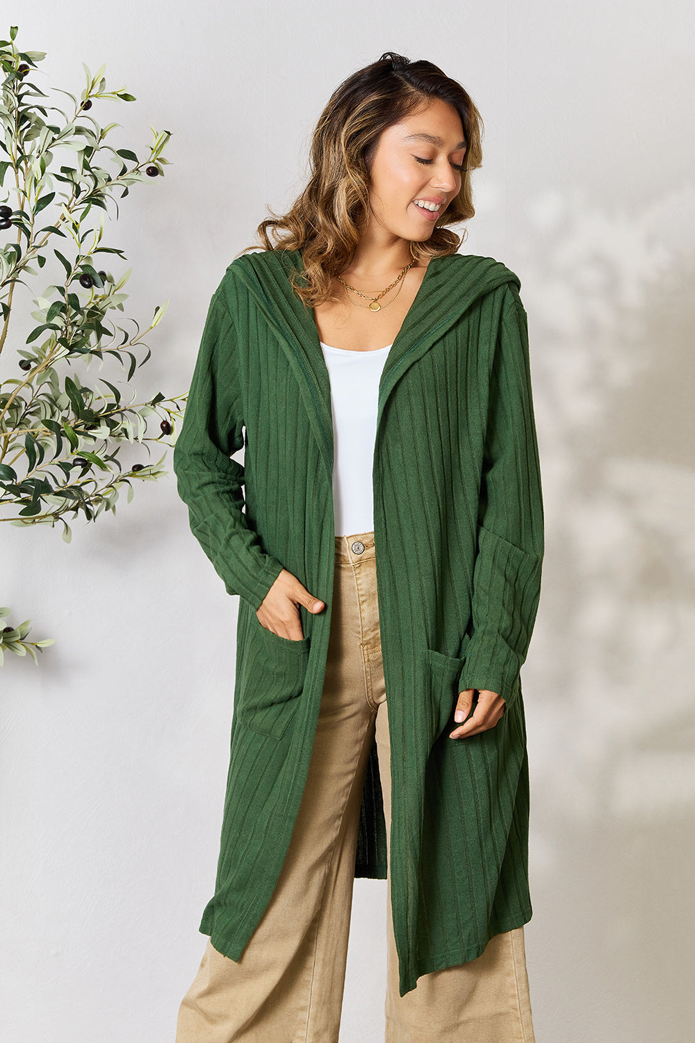 Woman wearing green hooded sweater cardigan with pockets, slightly stretchy fabric, styled with beige pants against a neutral background.