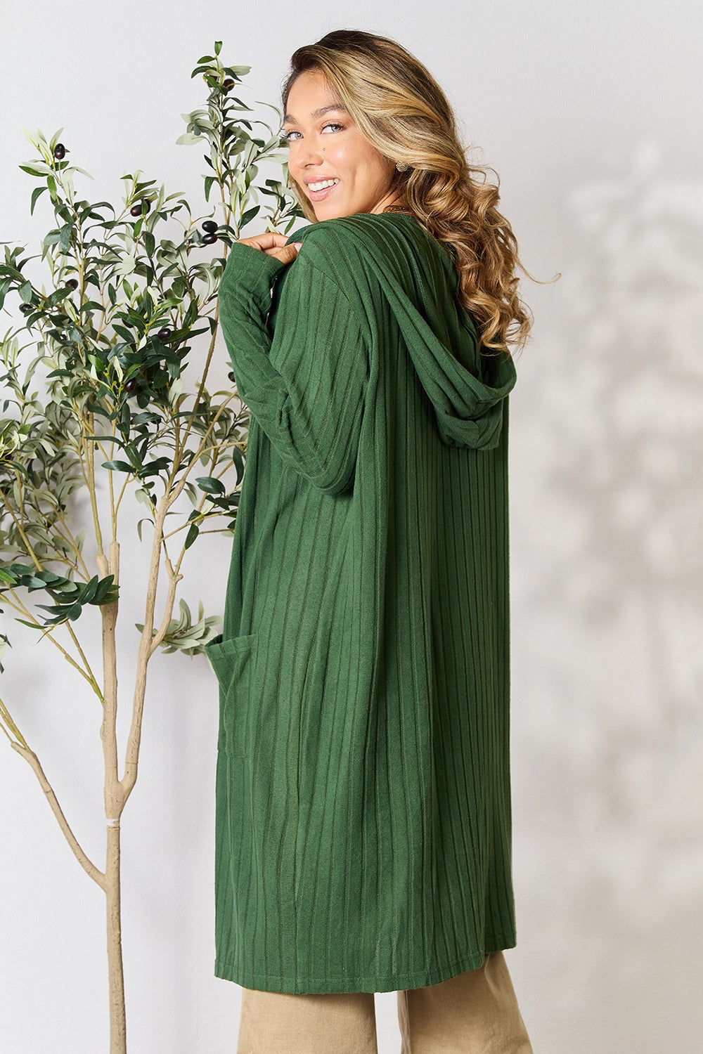 Woman wearing a green hooded sweater cardigan with pockets, standing next to an indoor plant, showcasing the back design.